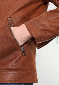 Giacca in pelle marrone con texture, dotata di tasche con zip e una finitura liscia, con dettagli in metallo argentato per resistenza e stile.