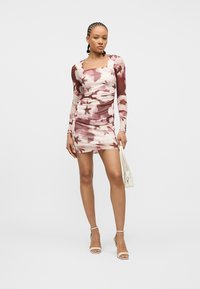 Γυναίκα με πλεξούδες, φορώντας ένα μίνι φόρεμα με μακριά μανίκια, ροζ και καφέ με λουλούδια, κρατώντας μια λευκή τσάντα και φορώντας λευκά σανδάλια με ψηλό τακούνι.