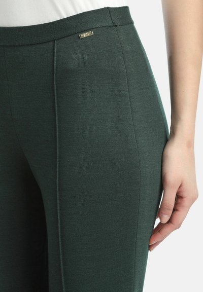 Tamsiai žali pritaikyti kelnės, pasižyminčios lygiu paviršiumi, subtilia siūlių detale ir mažu metaliniu logotipo akcentu ant juosmens.