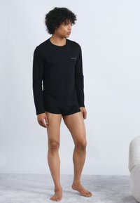 Jovem com cabelo encaracolado está descalço em cima de um tapete, vestindo uma camisa de manga longa preta e calções pretos, em frente a um fundo branco liso.