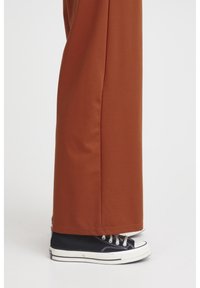 Pantalon à jambes larges de couleur rouille avec une texture lisse, associé à des baskets en toile noires avec des semelles blanches et un laçage classique.