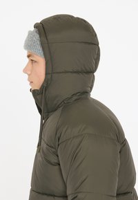 Olivenfarvet pufferjakke med hætte, med quiltet tekstur og snoretræk til udjævning. En grå strikket hue bæres under hætten.