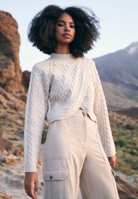 Lyscreme kabelstrikket sweater med cropped design, parret med beige bukser med brede ben, der har cargo-lommer og knapdetaljer.