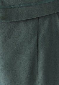 Ciemnozielony materiał o teksturowanym wykończeniu, z gładką talią i subtelnym detalem plisowania, co nadaje elegancki wygląd.
