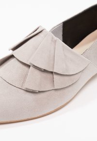 Lichtgrijze suède loafer met gelaagde franjerand bovenop en een gladde bruine zool, deels zichtbaar zwarte binnenvoering.