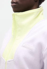 Κοντινό πλάνο ενός προσώπου που φοράει ένα fleece μπουφάν Columbia Sportswear σε ανοιχτό κίτρινο και λευκό χρώμα με ψηλό γιακά και κούμπωμα με φερμουάρ.