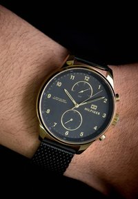 Montre dorée avec un cadran noir, indicateurs horaires numériques, deux sous-cadrans et un bracelet en maille noire texturé. Logo de la marque visible : Hilfiger.