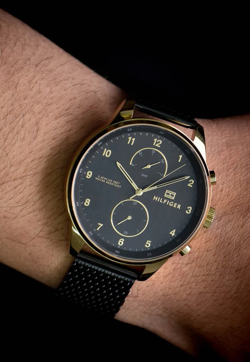 Montre dorée avec un cadran noir, indicateurs horaires numériques, deux sous-cadrans et un bracelet en maille noire texturé. Logo de la marque visible : Hilfiger.