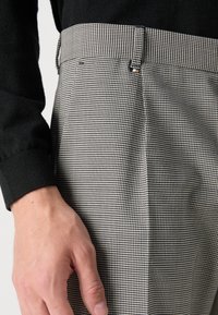 Detailné zobrazenie ruky odpočívajúcej pri bedrách, oblečenej v nohaviciach s čierno-bielym vzorom hounds-tooth a čiernej košeli s dlhým rukávom.