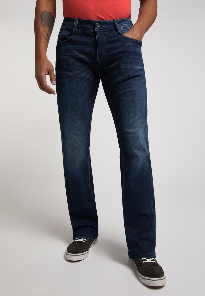 Homme portant un jean bleu foncé, des baskets noires et une chemise orange, avec une main dans la poche et un tatouage sur l'avant-bras gauche.