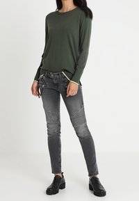 Mujer vestida con una blusa de manga larga de color verde oliva, jeans oscuros de estilo biker con detalles de cremalleras, y zapatos de plataforma negros y gruesos, de pie sobre un suelo blanco.