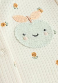 Lys ribbet cremefarvet stof har en grøn æblebrodering med et smilende ansigt, lyserøde kinder og ferskenfarvede blade, fremhævet af små blomstermotiver.
