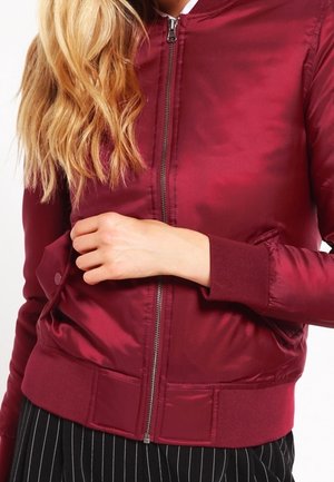 Femme portant une veste bomber bordeaux brillante avec fermeture éclair, poignets et taille côtelés, associée à un pantalon noir à fines rayures.
