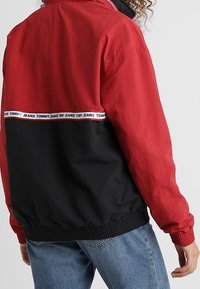 Osoba v červené a černé bundě Tommy Jeans s logem na pásce vzadu, v kombinaci s modrými džínami, otočená zády.