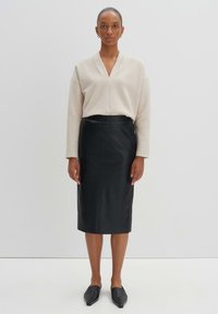 Maglione beige a scollo a V, veste largo, abbinato a una gonna a tubino nera in pelle, lunga fino al ginocchio. Mocassini neri a punta completano l'outfit.