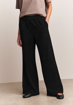 Persona che indossa pantaloni neri a gamba larga, una maglia taupe a maniche corte e scarpe basse nere, in piedi con una mano in tasca.