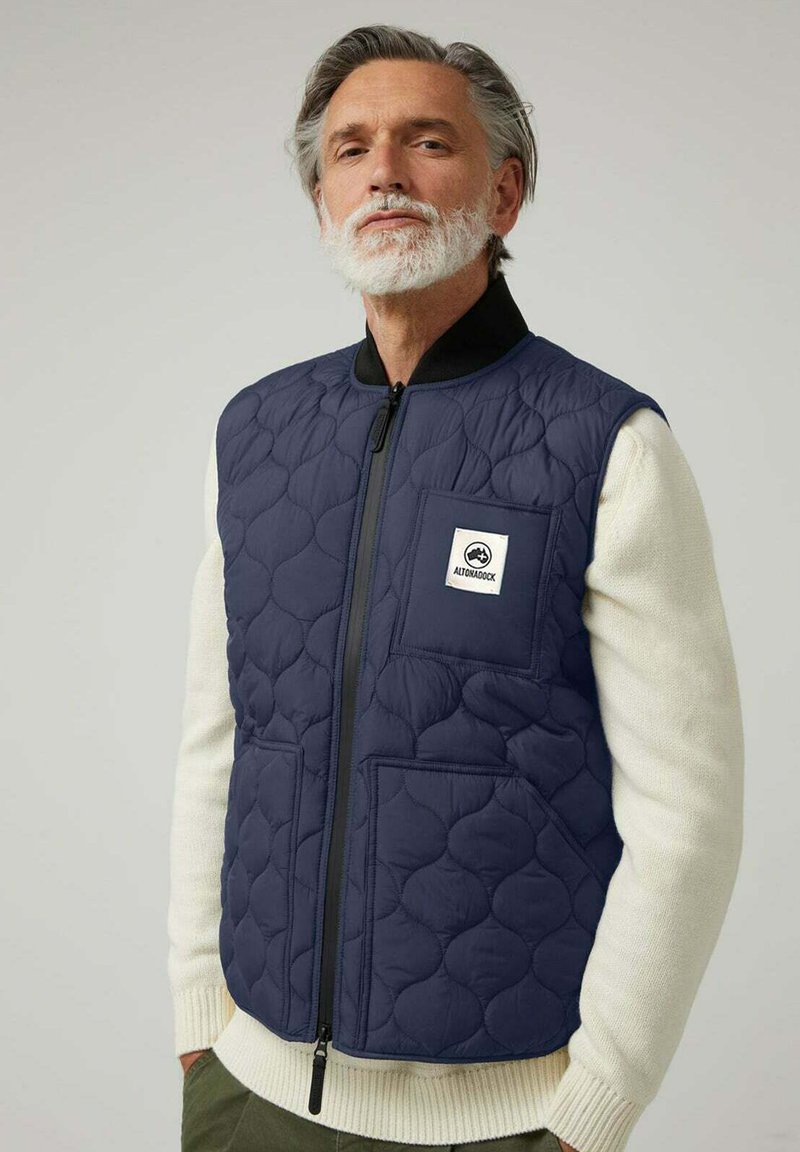 Chaleco acolchado color azul marino con cremallera frontal, textura acolchada y bolsillos laterales. Presenta un parche de logo blanco en el pecho izquierdo, llevado sobre un suéter beige.