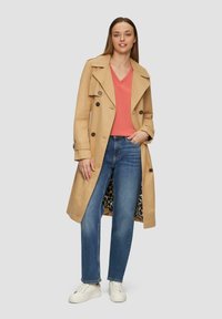 Beige trenchcoat met dubbele knoopsluiting, gedragen met een koraalkleurige V-hals top, blauwe jeans en witte sneakers; met zichtbaar leopardenprint binnenin.