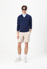 Laivastonsininen V-kaula-aukko villapaita vaaleansinisen aluspaidankin päällä, beige sävyinen raidallinen shortsit, valkoiset lenkkarit ja mustat aurinkolasit. Yksinkertainen, rento asu.