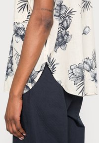 Blouse florale en crème avec motifs floraux et feuillages bleu marine, en tissu léger et doux, avec un ourlet arrondi et des fentes latérales, assortie à un pantalon sombre.
