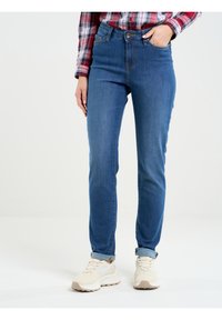 Augstas jostas zili džinsi ar pieguļošu formu, piedāvājot divas priekšējās kabatas un sarullētu apakšmalu, kombinēti ar gaišiem sporta apaviem.