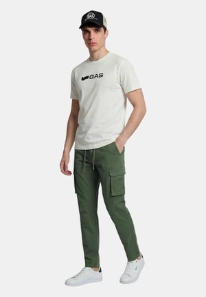 Giovane uomo con t-shirt bianca "GAS", pantaloni cargo verde oliva, sneakers bianchi e cappellino "GAS" nero e beige, in piedi con una mano in tasca.