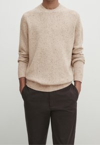 Vaalean beige neulepaita, jossa on pyöreä kaula-aukko, teksturoitu kangas, jossa on monivärisiä pilkkuja, ribbireunat hihoissa ja helmassa, yhdistettynä tummiin housuihin.