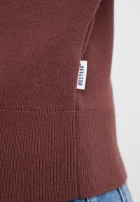 Burgunderfarbener Pullover aus feinem Strick mit geripptem Saum. Enthält ein kleines weißes Etikett mit "MUSTANG" in Blau, das an der Seite angebracht ist.