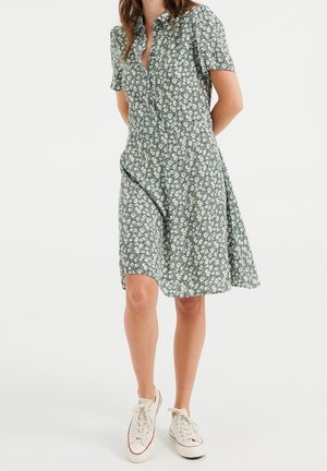 Kvinde iført en knælængde, kortærmet blomsterkjole med hvide sneakers, stående mod en ensfarvet hvid baggrund.