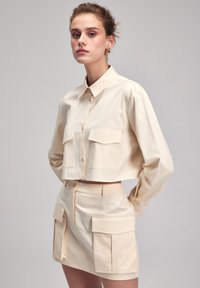 Korte beige blouse met twee borstzakken, knoopsluiting aan de voorkant en lange mouwen, gecombineerd met een bijpassende high-waisted rok met zijzakken.