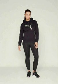 Sieviete, tērpusies melnā Puma kapucī, melnās legingās un melnās sporta kurpēs, stāv uz vienkārša gaiša fona.