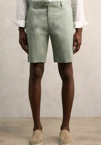 Pantaloni scurți din bumbac verde deschis, cu o croială ajustată, dotati cu un nasture frontal, buzunare laterale și o textură netedă. Purtați cu pantofi slip-on bej.