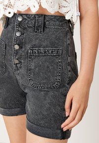 Shorts de mezclilla negros de tiro alto con botones frontales, puños enrollados, gran bolsillo delantero, combinados con un top de crochet blanco.