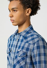 Camisa a cuadros azul y blanca con cuello abotonado, bolsillos en el pecho y una tela texturizada. Presenta costuras en contraste en los puños y las costuras.