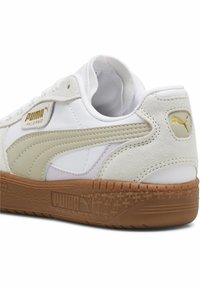 Baskets blanches avec une tige en suède et en cuir lisse, présentant une bande vert clair, des accents dorés et une semelle en caoutchouc gomme texturé.