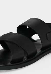 Μαύρο σανδάλι τύπου slide με δύο φαρδιές λουριά που διασταυρώνονται στο πόδι, μαλακή σόλα και λεία ματ επιφάνεια, εμφανίζεται σε απλό ανοιχτό φόντο.