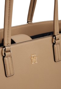 Μπεζ δερμάτινη τσάντα με υφή, διπλές λαβές και μικρό χρυσό λογότυπο TH στο μπροστινό μέρος, που δείχνει μέρος της σκουρόχρωμης εσωτερικής επένδυσης.