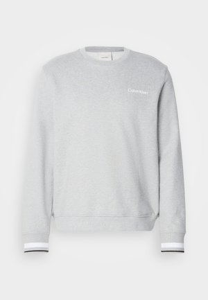 Svetlo siva majica sa dugim rukavima, napravljena od mekog materijala, sa okruglim izrezom, rebrastim manžetama sa crnim i belim prugama, i malim logotipom na grudima.