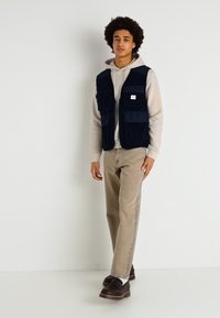 Gilet in tessuto navy con molte tasche sopra un leggero hoodie, abbinato a pantaloni beige e zoccoli scuri, in piedi davanti a uno sfondo bianco.