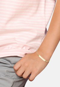 Pulsera de cadena de oro con una etiqueta rectangular en blanco para grabar, llevada en una muñeca, sobre una camisa a rayas rosas.