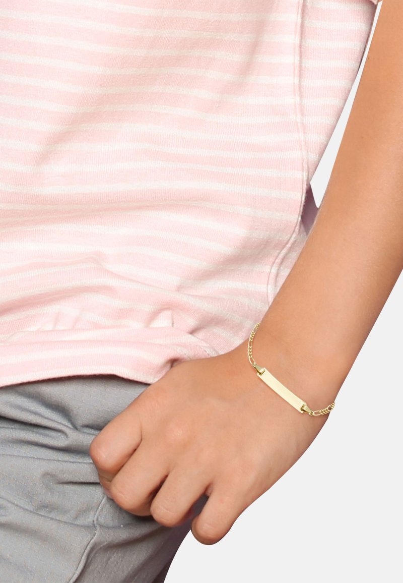 Pulsera de cadena de oro con una etiqueta rectangular en blanco para grabar, llevada en una muñeca, sobre una camisa a rayas rosas.