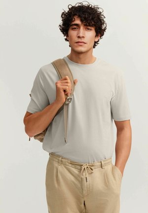 Junger Mann mit lockigem Haar, der ein hellgraues T-Shirt und beige Hose trägt und einen hellbraunen Rucksack über einer Schulter trägt.