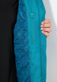 Teal pufferjacka med en slät ytteryta och quiltad innervadding, med en dragkedjeficka och mönster med texten "HYRAR".