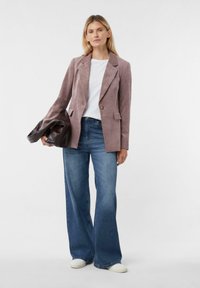 Lichtroze corduroy blazer met notch-lapels, gecombineerd met een wit T-shirt en wijde blauwe jeans. Houdt een donkerbruine leren tas vast.