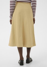 Une jupe midi beige en coton avec une texture lisse, présentant des détails de couture et une légère évasement à l'ourlet. Des chaussures noires complètent le look.