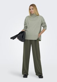 Suéter de gran tamaño de color verde claro con cuello alto y dobladillo acanalado, combinado con pantalones holgados en un tono verde oliva y un bolso negro. Textura suave, ajuste relajado.