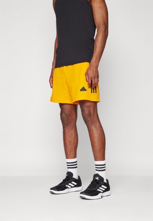 Schwarzes ärmelloses Tanktop, gelbe Shorts mit schwarzen Streifen und Logo, schwarze Sportschuhe und weiße Socken mit schwarzen Streifen.