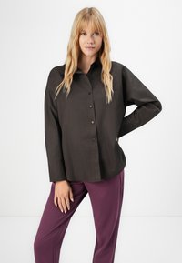 Blond kvinne iført en løs mørk skjorte med knapper og lilla bukser, står med den ene hånden på hoften og et nøytralt uttrykk.