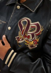 Blouson en cuir noir avec un grand motif brodé aux éléments flamboyants et aux initiales "PR". Poignets et col côtelés à rayures.