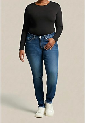 Kvinde iført mørkeblå jeans, en tætsiddende sort langærmet top og hvide sneakers, stående mod en ensartet lys baggrund.